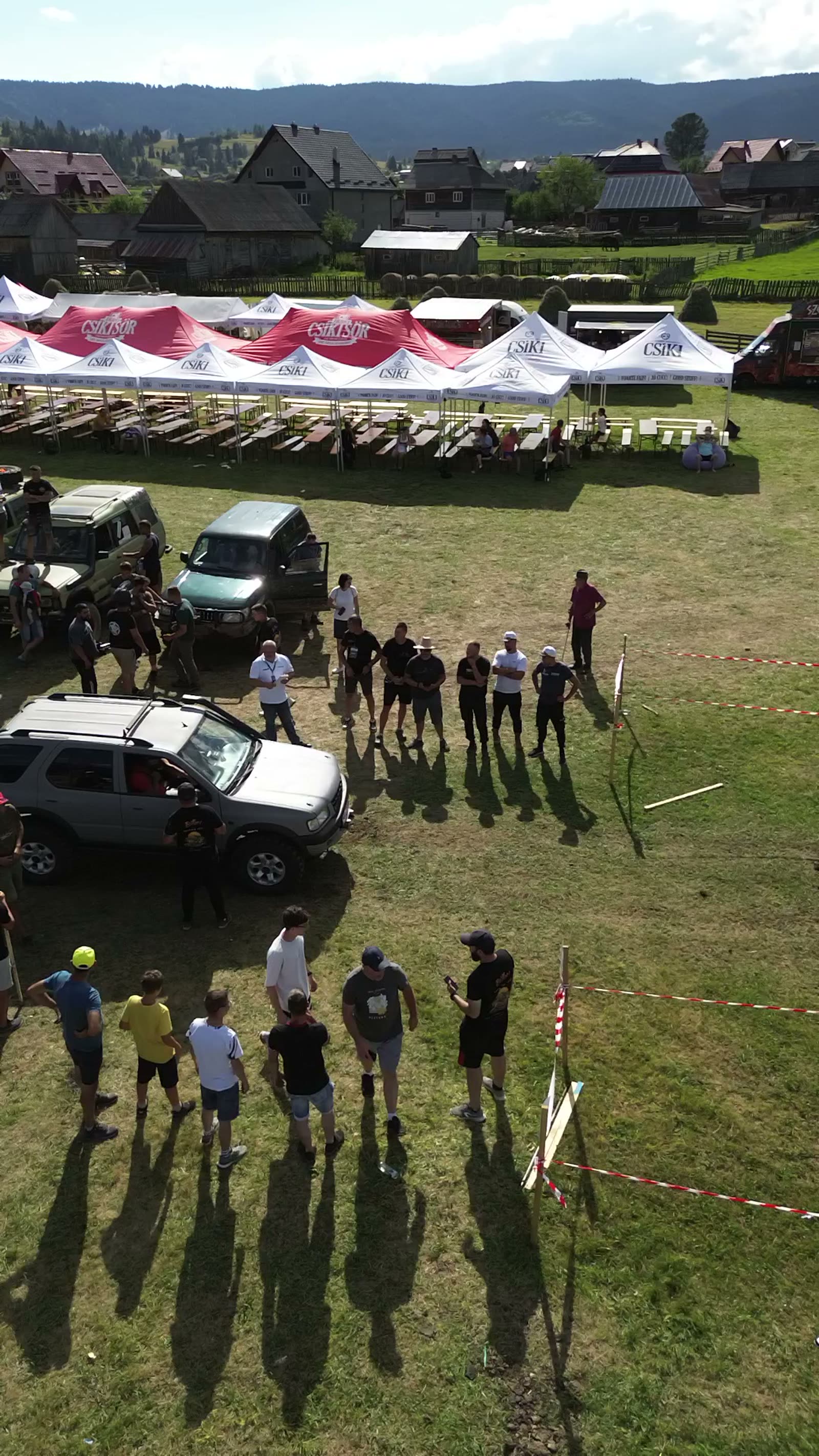 Vedere de sus la Bilbor Offroad Fest: mașini 4x4 și echipaje adunate pe iarbă lângă banda roșu-albă, cu corturi albe și roșii, mese și case de lemn în fundal